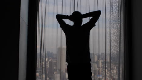 Silhouette-of-a-man-stretching-and-looking-out-the-window-with-the-curtains-closed,-obscuring-the-panoramic-view-of-the-city-metropolis