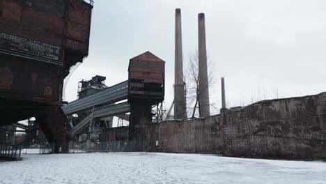 Old,-abandoned-industrial-complex-covered-in-snow,-with-rusted-structures,-buildings-and-tall-chimneys-standing-against-an-overcast-sky