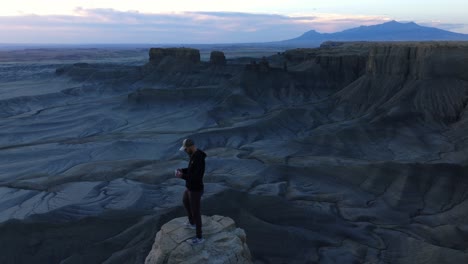 Mann-Steht-Auf-Einem-Felsen-Und-Steuert-Eine-Drohne-über-Die-Unglaubliche-Mondähnliche-Landschaft-Des-Factory-Butte-In-Utah,-USA
