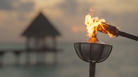 Steel-oil-lamp-on-post-being-lit-by-burning-torch-with-dramatic-sunset-in-background