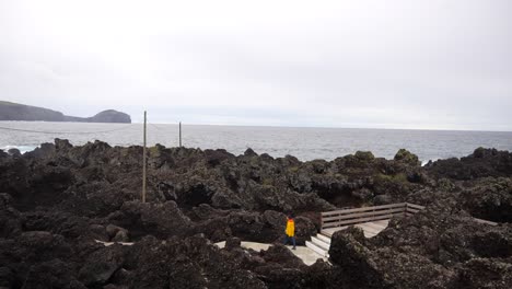 Young-man-in-yellow-Jjcket-walking-in-a-path-between-volcanic-rocks-on-Varadouro-Natural-Pools,-Faial