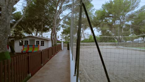 A-drone-glides-parallel-to-a-children's-playground-adorned-with-miniature-houses-and-a-tree-park,-and-along-a-field-for-both-volleyball-and-football-in-the-vibrant-locale-of-Mallorca,-Spain