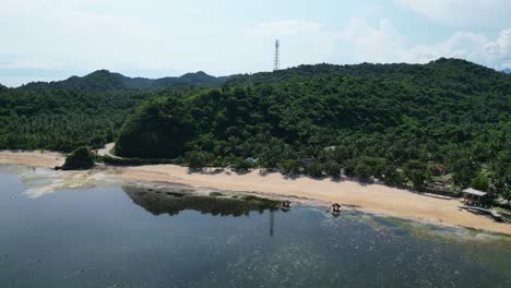 Aerial-drone-shot-of-Philippine-beach-resort-shoreline-with-lush-mountains-and-satellite-tower-in-background
