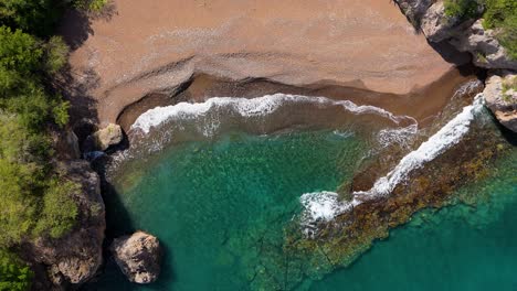 Drohne-Erhebt-Sich-über-Dem-Schwarzen-Sandstrand-Von-Playa-Santu-Pretu-Mit-Meereswellen,-Die-Auf-Felsplatten-Krachen