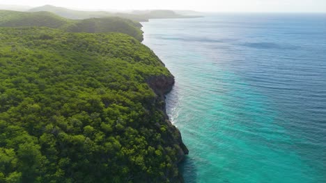 Lush-green-tropical-cliffs-spill-into-sandy-coral-reefs-and-drop-off-into-deep-blue-ocean,-aerial-dolly-split-land-and-water