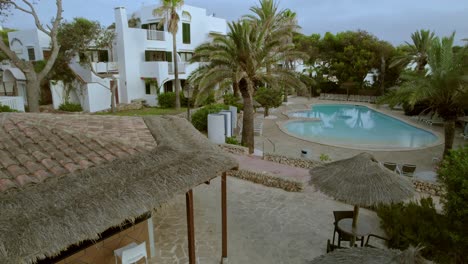 Luftaufnahme-Eines-Touristenresorts-Auf-Mallorca,-Spanien,-Mit-Einem-Unterkunftsgebäude-Mit-Einem-Mit-Stühlen-Und-Tischen-Geschmückten-Vorgarten-Und-Einem-Offenen-Swimmingpool