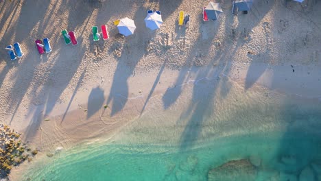 Crystal-clear-ocean-waves-crash-gently-on-golden-sand-beach-with-bright-colored-chairs-and-umbrellas,-aerial-top-down