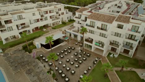Aerial-view-of-a-tourist-resort-in-Mallorca,-Spain,-featuring-a-three-story-accommodation-building-with-a-front-yard-adorned-with-chairs-and-tables