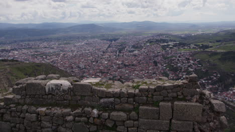 Un-Antiguo-Muro-De-Piedra-Con-Una-Ciudad-Y-Montañas-Al-Fondo-En-Pérgamo