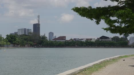 City-skyline-viewed-across-a-tranquil-lake-with-greenery,-under-a-clear-sky,-daytime,-urban-peacefulness