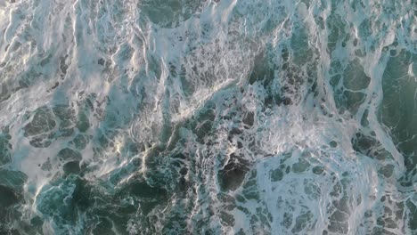 Rising-aerial-view-looking-straight-down-onto-breaking-waves-as-swells-come-in-creating-ebb-and-flow-movement-with-white-wash-giving-a-sense-turbulence