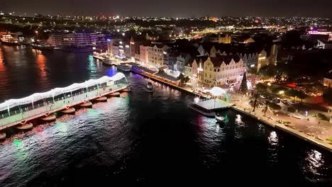 Tug-boat-exits-around-Queen-Emma-pontoon-bridge-lit-up-at-night,-aerial
