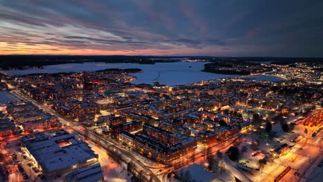 Luleå-city-at-dusk-with-city-lights-reflecting-on-snowy-streets-and-frozen-waterways,-winter-season,-aerial-view