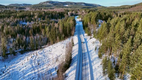 Car-on-snowy-road-amidst-pine-forest-in-Skorped,-Sweden,-with-shadows-stretching-across-the-landscape,-aerial-view