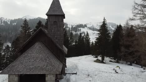 revealing-shot-old-stone-and-wood-church-in-winter-forest-with-a-traveller-showing-a-ski-resort-with-hotels-and-slopes