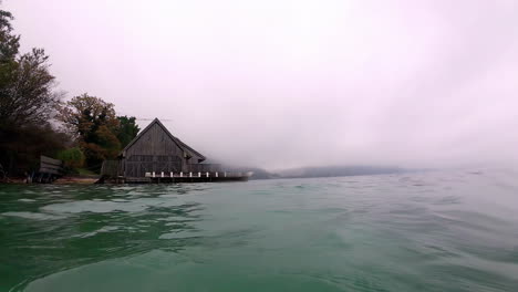 Wooden-hut-on-lakeside-with-mist-covered-mountains-in-distance