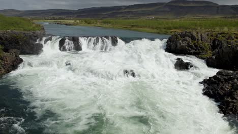 Ein-Fluss-In-Island,-Der-Stark-über-Felsen-Fließt,-Mit-Hügeln-Und-Wolken-Im-Hintergrund