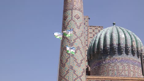 Uzbek-flags-flying-in-front-of-blue-tiled-domes-and-minaret-in-Samarkand,-Uzbekistan-along-the-historic-Silk-Road
