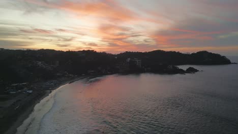 Drone-rises-above-surfers-catching-rolling-waves-into-sandy-bay-at-sunset,-Sayulita-Mexico