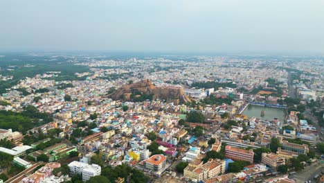 Malaikottai-rock-fort-in-middle-of-Tiruchirappalli-city's-dense-urban-buildings,-aerial-panoramic-dolly