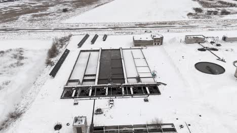 Drone-flies-over-water-treatment-plant-located-in-small-towns-in-winter,-snow-covered-landscape