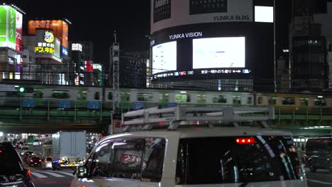 Evening-Traffic-Driving-Past-In-Shinjuku-With-Local-Metro-Train-Seen-Going-Across-On-Elevated-Railway-Tracks-Show-Urban-Charm-In-Tokyo-Metropolis