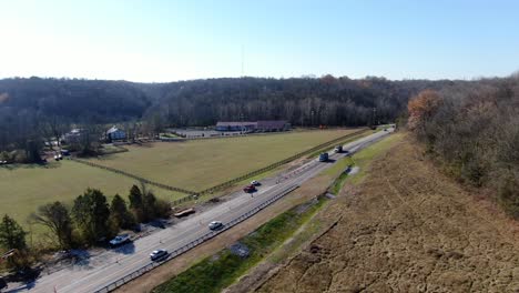 Aerial-Shot-of-Cars-Driving-Along-a-Rural-Highway-Between-Some-Fields-and-Forests-on-a-Sunny,-Winter-Day