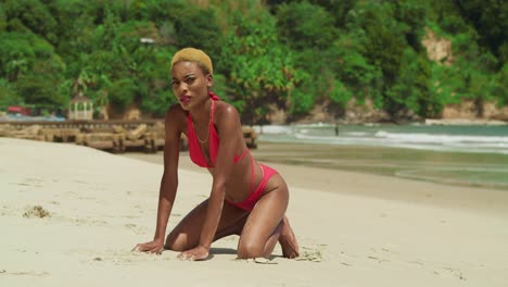 On-the-pristine-shores-of-a-Caribbean-beach,-a-girl-with-dark-skin-stands-out-in-a-red-bikini-kneel-in-the-sand