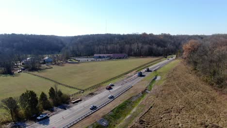 Aerial-Shot-of-Cars-Driving-Along-a-Rural-Highway-Between-Some-Fields-and-Forests-on-a-Sunny,-Winter-Day