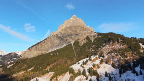 Sunlit-rocky-peak-in-Alps-mountains-with-snowy-forest-mountainside