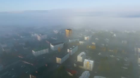 Aerial-view-circling-a-cityscape-covered-in-thick-mist,-autumn-sunrise-in-Scandinavia