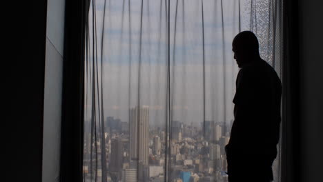 A-man's-silhouette-paces-back-and-forth,-gazing-out-a-window-with-closed-curtains,-concealing-the-expansive-city-metropolis-view