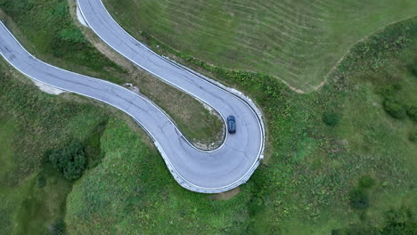 Car-taking-sharp-curving-turn-on-grassy-woodland-mountain-road-aerial-top-down-view