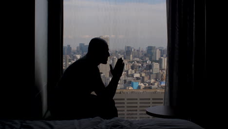 Silhouette-Of-Lonely-Male-Anxiously-Sitting-Near-High-Rise-Apartment-Window,-Rocking-Back-Showing-Loneliness,-Thinking-And-Anxiety