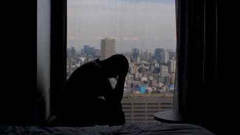 silhouette-of-adult-man-anxiously-sitting-near-high-rise-window,-shaking-his-head-in-his-hands-with-panoramic-view-of-the-city-metropolis-in-background