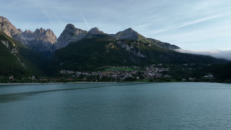 Drone-flying-over-lake-with-Molveno-village-and-Dolomites-mountains-in-background,-Trentino-in-Italy