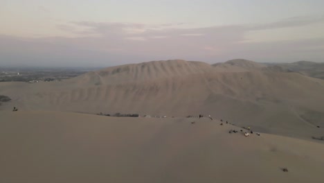 Drone-gracefully-soars-over-the-dunes,-passing-by-people,-revealing-the-oasis-gem,-Huacachina