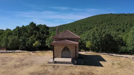 flight-with-close-up-of-a-hermitage-and-when-moving-away-you-see-that-it-is-in-a-meadow-with-a-pond-surrounded-by-a-road-in-a-forest-with-the-valley-in-the-background-in-Avila,-Spain