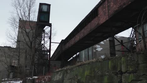 Old,-dilapidated-industrial-building-with-a-rusted-bridge-connecting-two-sections,-surrounded-by-overgrown-vegetation-under-a-cloudy-sky