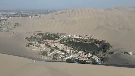 Experience-the-allure-of-Huacachina-as-the-drone-gracefully-moves-forward,-showcasing-its-unique-blend-of-tranquil-lagoon-and-majestic-dunes—an-aerial-journey-through-the-heart-of-Peru's-desert-oasis