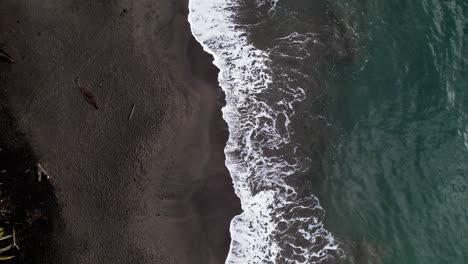 Caribbean-water-washing-volcanic-dark-beach-of-Guadeloupe,-aerial-top-down