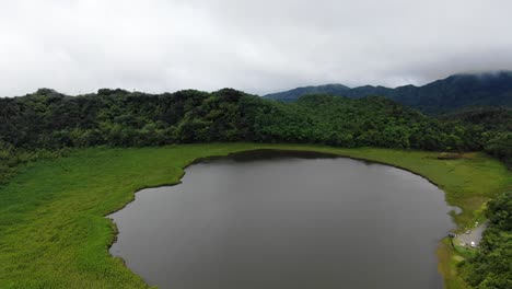 Volcanic-crater-lake-reflects-cloudy-sky-in-Grand-Etang-National-Park,-Aerial-panoramic-establish