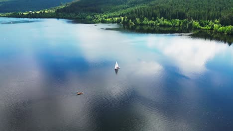 A-sailboat-on-a-serene-lake-in-the-black-forest,-Germany,-surrounded-by-lush-greenery,-reflecting-clouds,-aerial-view