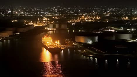Oil-tanker-docked-at-refinery-reflects-shimmering-yellow-light-on-water,-aerial