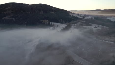 Aerial-view-of-landscape-with-a-forested-hill,-open-fields,-and-low-lying-fog,-creating-a-mystical-atmosphere