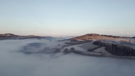 Aerial-view-of-tranquil-landscape-at-dawn,-with-rolling-hills-emerging-from-a-thick-layer-of-mist,-bathed-in-soft-sunlight