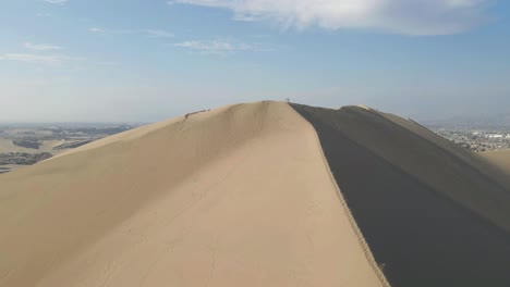 Drone-ascends-the-Ica-dunes,-unveiling-the-breathtaking-panorama-beyond-the-peak