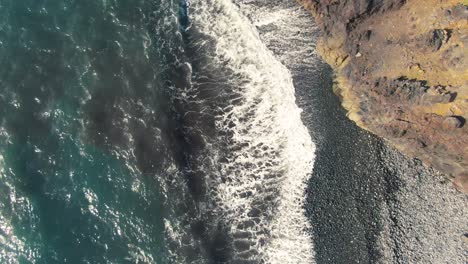 Ocean-waves-wash-over-stone-beach-by-cliff-in-Hawaii,-overhead-aerial