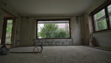 Wide-shot-on-a-construction-site,-living-room-gets-renovated,-camera-tracks-towards-a-huge-window,-beautiful-garden-with-bushes-visible