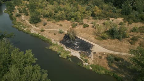 Aerial-revealing-opening-shot-of-burned-dry-grass-by-the-river-on-hot-summer-day,-Europe